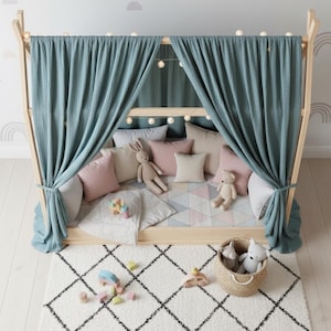 May include: A wooden playhouse bed with a teal curtain, string lights, and various pillows in pink, beige, and gray. A white and black patterned rug is in front of the bed, with a basket of stuffed animals and wooden toys.