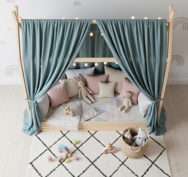 May include: A wooden playhouse bed with a teal curtain, string lights, and various pillows in pink, beige, and gray. A white and black patterned rug is in front of the bed, with a basket of stuffed animals and wooden toys.