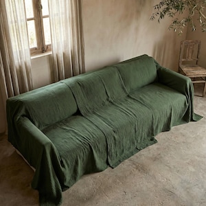 May include: A green sofa draped with a textured throw. The room features a window with curtains, a wooden chair, and a neutral-toned wall. The scene evokes a sense of calm and simplicity.