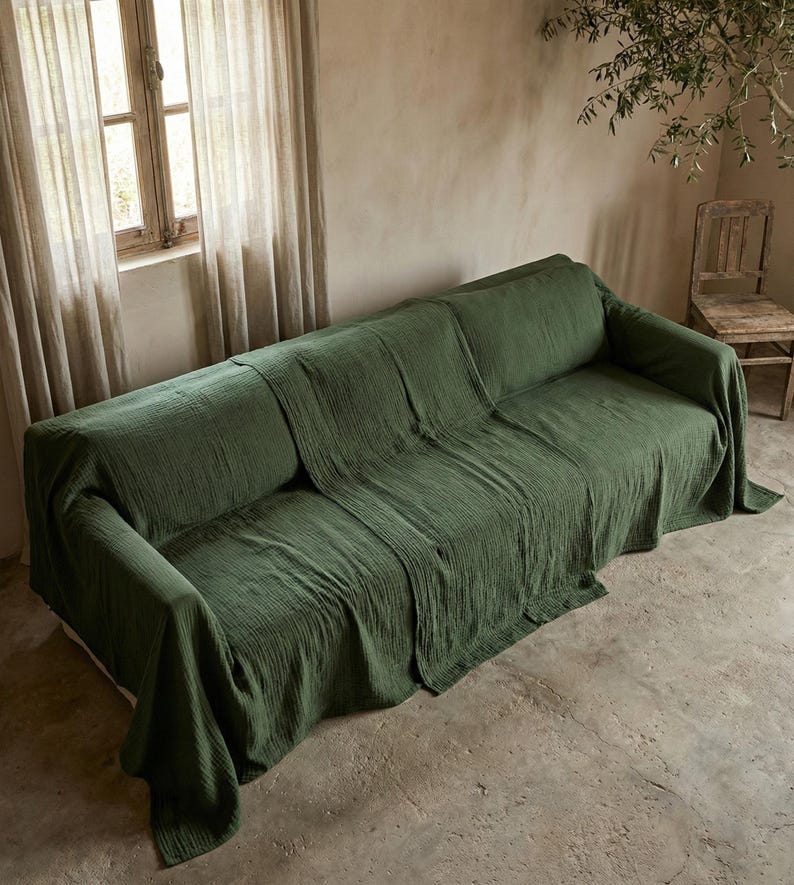 May include: A green sofa draped with a textured throw. The room features a window with curtains, a wooden chair, and a neutral-toned wall. The scene evokes a sense of calm and simplicity.
