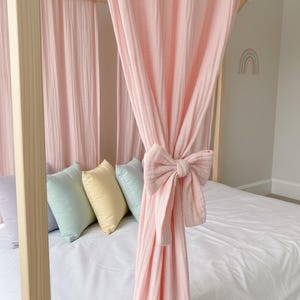 May include: A light pink muslin canopy curtain draped over a wooden bed frame, featuring a matching fabric bow tie-back. The bed is dressed with a white sheet and a row of four small square pillows in lavender, mint green, yellow, and light blue.