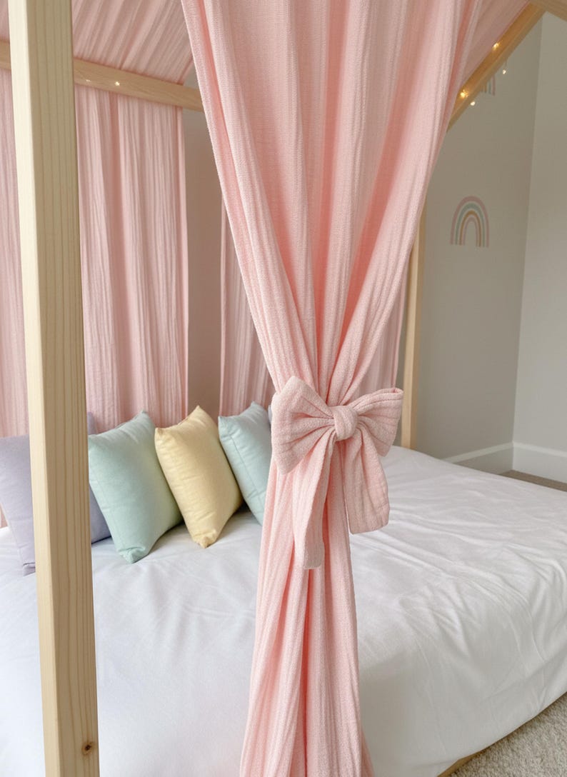 May include: A light pink muslin canopy curtain draped over a wooden bed frame, featuring a matching fabric bow tie-back. The bed is dressed with a white sheet and a row of four small square pillows in lavender, mint green, yellow, and light blue.