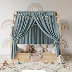 May include: A wooden house-shaped bed frame with a light blue curtain and string lights. The bed is decorated with pillows, a teddy bear, and a blanket. A white rug and toys are in front of the bed.