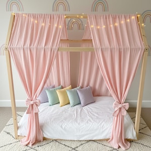 May include: A wooden house-shaped bed frame draped with soft pink fabric curtains tied with bows. The bed features a white mattress, five small square pillows in pastel shades of lavender, mint, yellow, and blue, and a string of warm fairy lights.