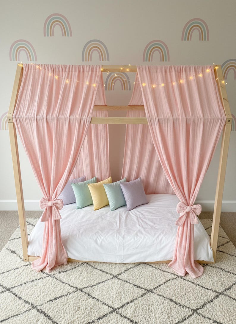 May include: A wooden house-shaped bed frame draped with soft pink fabric curtains tied with bows. The bed features a white mattress, five small square pillows in pastel shades of lavender, mint, yellow, and blue, and a string of warm fairy lights.
