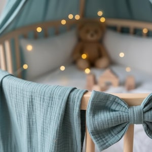 May include: A baby crib with a soft blue blanket draped over the side. A large bow is attached to the crib. A teddy bear and wooden blocks are inside the crib, with string lights adding a warm glow.