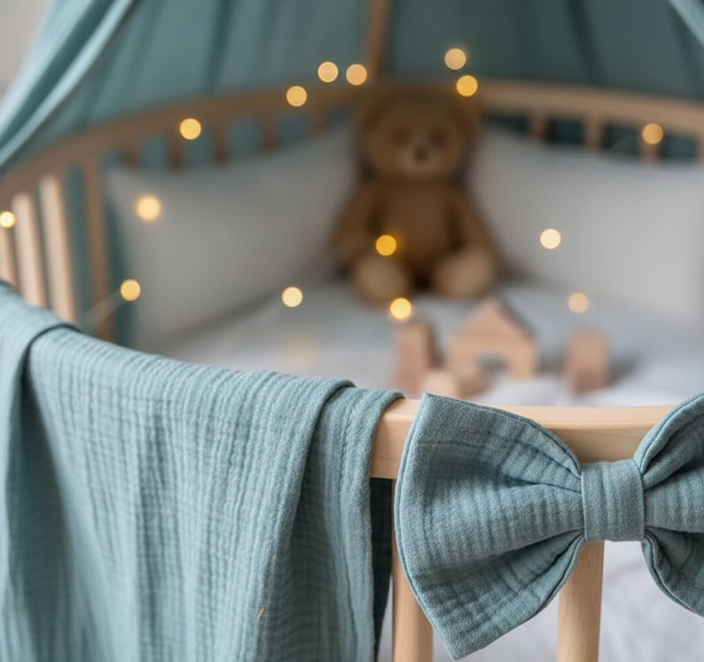 May include: A baby crib with a soft blue blanket draped over the side. A large bow is attached to the crib. A teddy bear and wooden blocks are inside the crib, with string lights adding a warm glow.