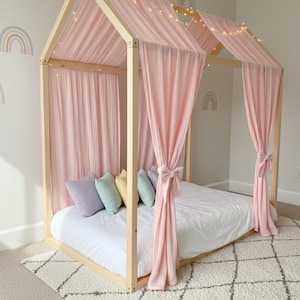 May include: A light-colored wooden house-shaped bed frame draped with soft pink fabric curtains tied at the sides. The bed features a white mattress, a row of pastel-colored throw pillows in lavender, mint, yellow, and blue, and string lights.