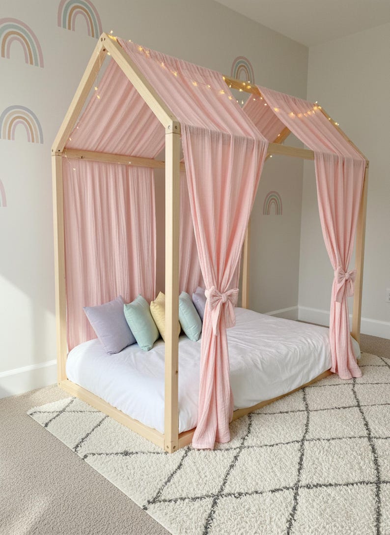 May include: A light-colored wooden house-shaped bed frame draped with soft pink fabric curtains tied at the sides. The bed features a white mattress, a row of pastel-colored throw pillows in lavender, mint, yellow, and blue, and string lights.