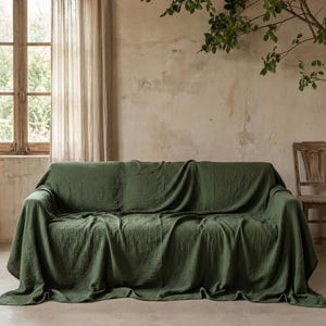 May include: A green sofa throw draped over a sofa. The throw is a solid, muted green colour and has a textured appearance. The sofa is positioned in front of a neutral-toned wall, with a window and a wooden chair visible in the background.
