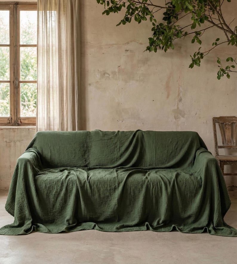 May include: A green sofa throw draped over a sofa. The throw is a solid, muted green colour and has a textured appearance. The sofa is positioned in front of a neutral-toned wall, with a window and a wooden chair visible in the background.