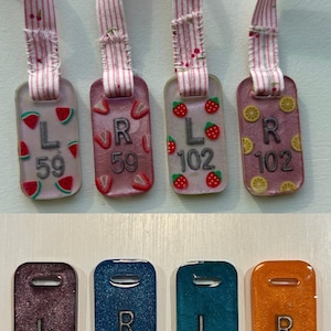 May include: Luggage tags in various colours and designs. Some have fruit illustrations, including watermelon, strawberries, and lemons. Others are solid colours with the letters "L" or "R" and numbers. Each tag has a striped ribbon.