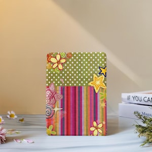 May include: A rectangular decorative plaque featuring a split design. The top half is olive green with a white star pattern, while the bottom half has vertical stripes in pink, orange, yellow, and purple. It is decorated with yellow flowers, stars, and a pink butterfly.