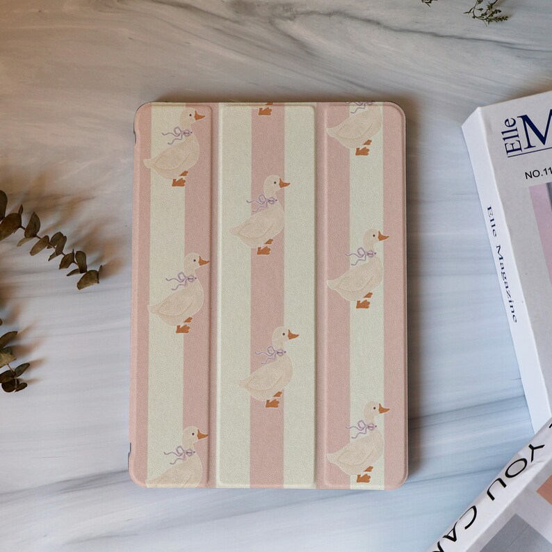 May include: A pink and white striped tablet case featuring a pattern of cartoon geese with lavender bows. The case is closed and has a magnetic cover. The case is next to a white book with the word "Elle" on the spine.