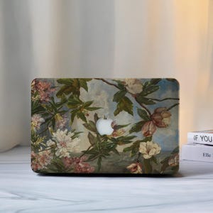 Peut inclure: Coque d'ordinateur portable avec un motif floral. Le design présente des fleurs roses, blanches et crème, des feuilles vertes et un fond bleu clair. Elle possède un logo Apple blanc.
