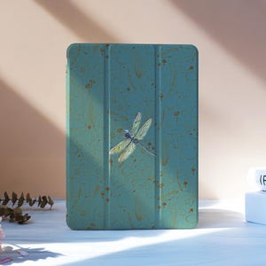 Puede incluir: Funda de tableta turquesa con una ilustración de libélula. La funda tiene un aspecto texturizado y veteado con detalles dorados. La libélula tiene alas detalladas y un cuerpo delgado. La funda está sobre una superficie blanca.