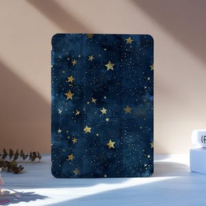 Può includere: Una custodia per tablet blu scuro con un design celeste con stelle dorate e minuscoli puntini bianchi. La custodia è in piedi su una superficie bianca, con due libri sullo sfondo. La custodia ha angoli arrotondati.