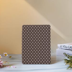 May include: A brown tablet case with a white polka dot pattern. The case is rectangular and stands upright on a white surface. The background is a light brown color.