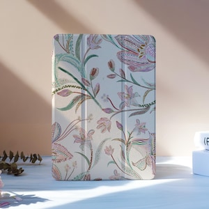 Könnte beinhalten: Eine Tablet-Hülle mit floralem Muster auf cremefarbenem Hintergrund, mit rosa, grünen und braunen botanischen Designs. Die Hülle steht auf einer hellblauen Oberfläche, mit Büchern im Hintergrund.