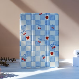 May include: A tablet case with a blue and white chequered pattern, decorated with red hearts and cherries. The case is on a white surface, with flowers and books in the background.