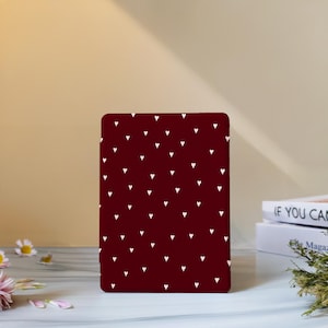 Può includere: Un oggetto rettangolare, color bordeaux, con un motivo di piccoli cuori bianchi distanziati in modo uniforme. L'oggetto è in posizione verticale su una superficie bianca, con uno sfondo sfocato con libri e fiori.