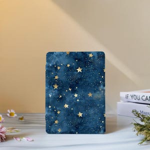 May include: A rectangular navy blue case with a celestial design. It has a watercolour-style background with gold stars and tiny white dots. The rounded corners give it a modern look. The case is likely for a tablet or e-reader.