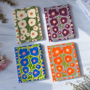 May include: Four rectangular notebooks featuring floral patterns. One has white flowers on green, one has purple flowers on a dark background, one has blue flowers on a yellow background, and one has orange flowers on a light blue background.
