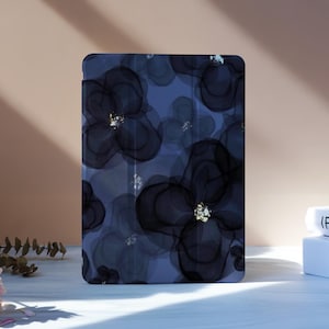 May include: A tablet case with a dark blue floral pattern, black and gold details. The case is rectangular and upright. The background shows a light surface and two books. The case's dimensions are approximately 20 cm x 15 cm.