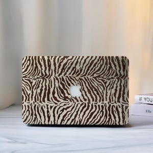 Può includere: Una custodia per laptop con un design a stampa zebrata. La custodia presenta un motivo a strisce marroni su sfondo crema. Il logo Apple è visibile al centro.