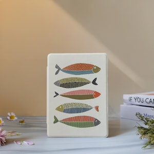 Puede incluir: Una funda rectangular blanquecina con cinco ilustraciones de peces estilizados en varios colores. Los diseños de los peces incluyen naranja, verde, azul y rosa, con patrones detallados. La funda está de pie.