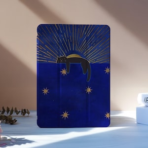 May include: A blue tablet case featuring a black cat lounging against a gold sunburst design. Gold stars are scattered across the blue background. The case is on a white surface with flowers and books in the background.