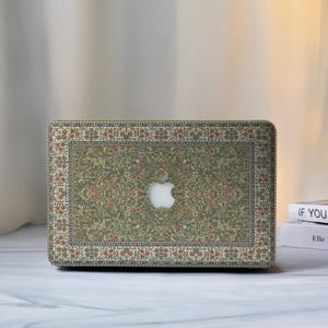 Puede incluir: Funda para portátil con diseño de alfombra. La funda tiene un fondo verde con patrones florales y geométricos en rojo, naranja y crema. El logotipo de Apple es visible. La funda está diseñada para un portátil.