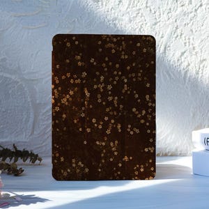 Puede incluir: Una funda rectangular para tableta en acabado marrón oscuro con un patrón disperso de pequeñas flores de cerezo de color dorado en toda la superficie. La funda tiene un panel frontal liso y plano con esquinas redondeadas y orientación vertical.