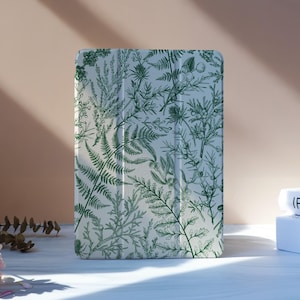 Op de afbeelding: Een tablethoesje met een botanisch ontwerp met groene varen- en bladerillustraties op een witte achtergrond. De hoes staat rechtop op een wit oppervlak, met roze bloemen en boeken op de achtergrond.