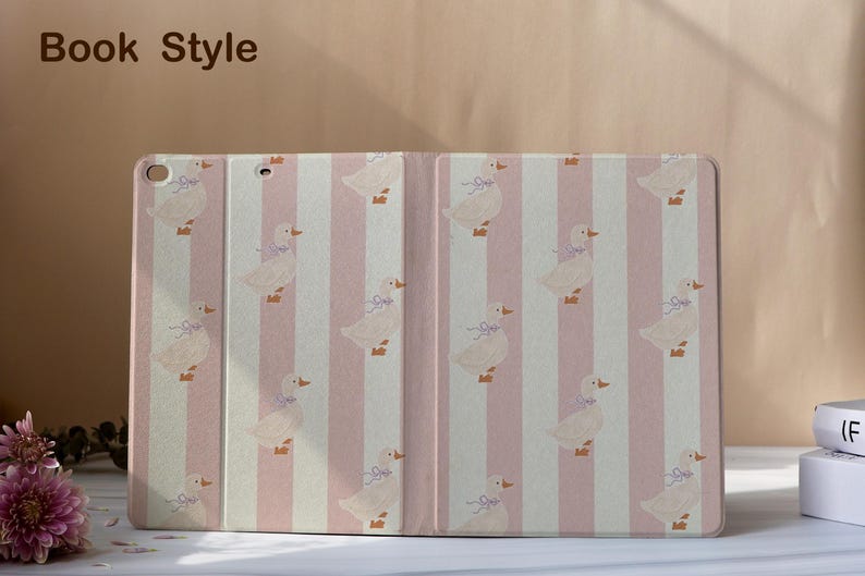 May include: A tablet case with a pink and white striped pattern. The case features a repeating pattern of ducks with blue bows. The words "Book Style" are in the upper left corner.