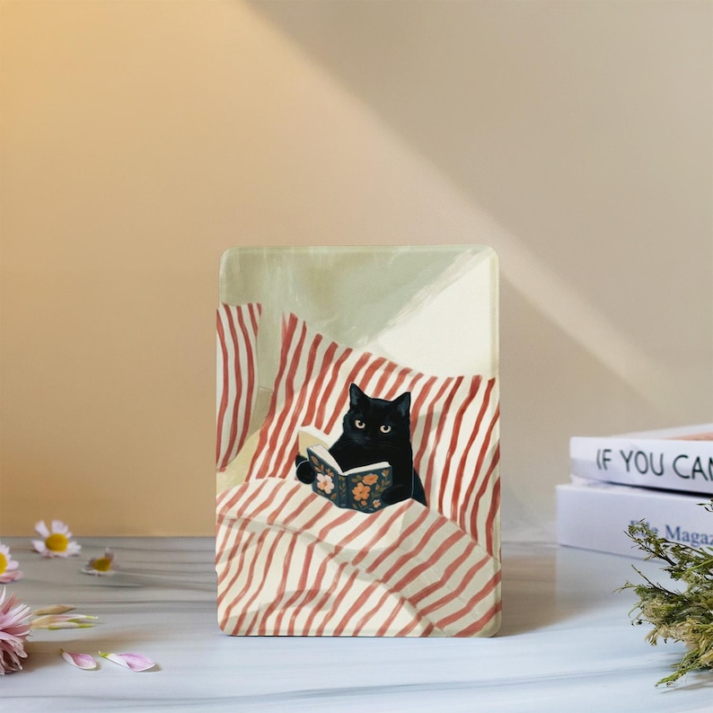 May include: A rectangular print featuring a black cat sitting in bed with red and white striped bedding, holding a small open book with a floral cover. The cat has bright yellow eyes and is positioned against a soft, light-coloured background.