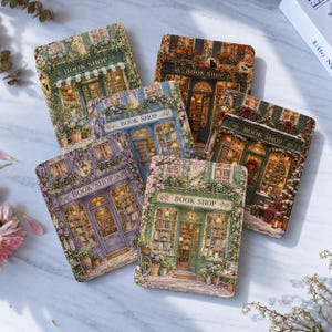 Op de afbeelding: Een set van vijf rechthoekige onderzetters met gedetailleerde illustraties van boekwinkels in verschillende seizoenthema's, elk met de tekst BOOK SHOP boven de ingang en voorzien van diverse florale en architecturale details.