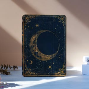 Puede incluir: Una cubierta de libro azul oscuro con un diseño de luna creciente dorada y estrellas. La cubierta tiene detalles dorados ornamentados en los bordes. El libro está de pie sobre una superficie blanca, con flores y otros libros en el fondo.