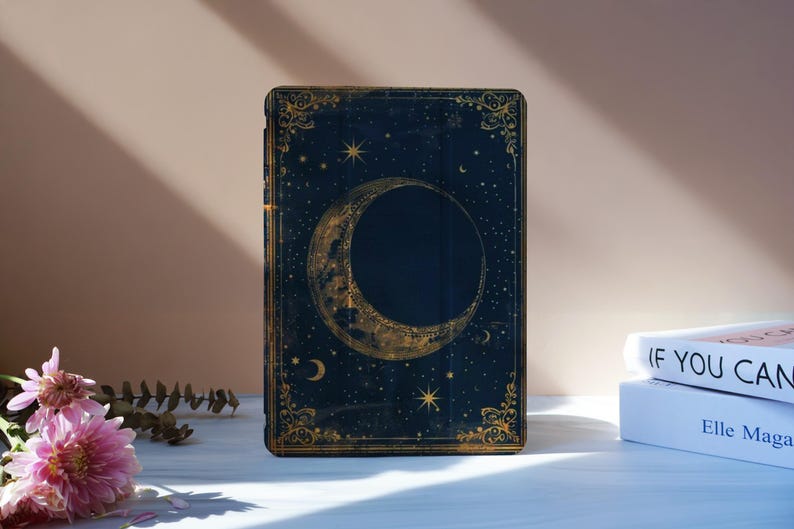 May include: A dark blue book cover with a gold crescent moon and stars design. The cover has gold ornate detailing around the edges. The book is standing upright on a white surface, with flowers and other books in the background.