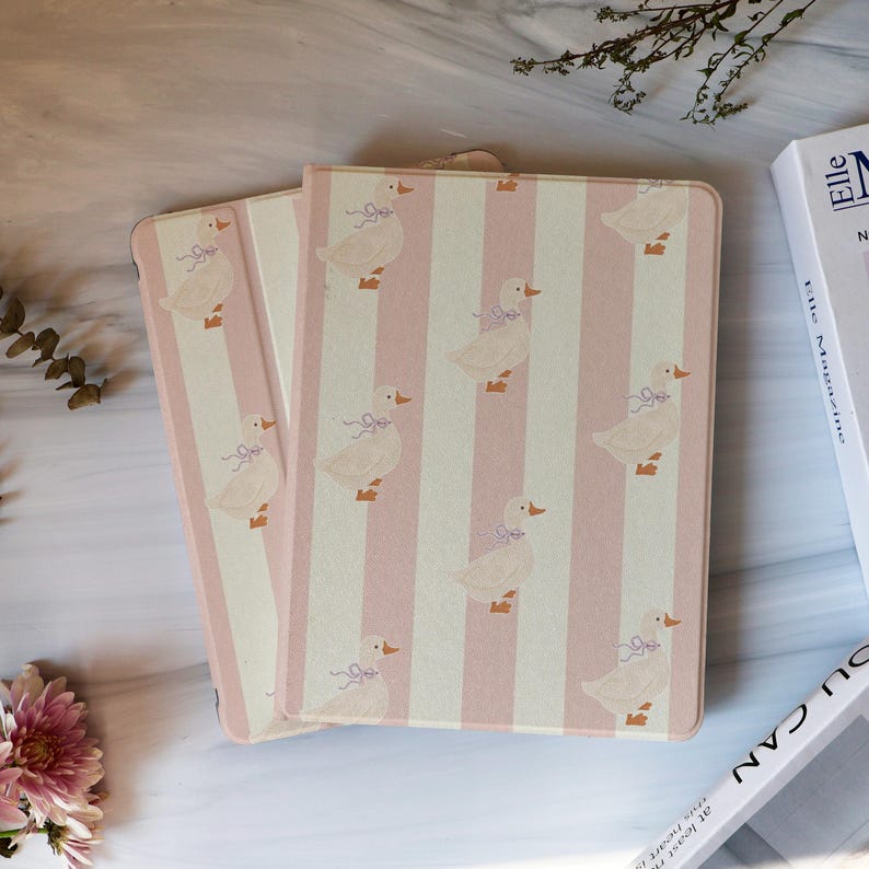 May include: Two pink and white striped notebooks with a pattern of white ducks wearing purple bows. The notebooks are rectangular and have a soft, pastel color scheme. The notebooks are closed and lying next to each other.