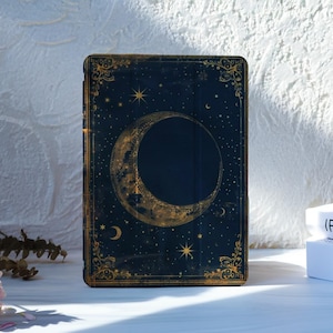 May include: A dark blue book cover with a gold crescent moon and stars design. The cover has a gold border with ornate detailing. The book is standing upright on a white surface, with a stack of books in the background.
