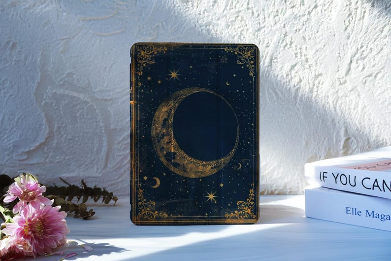 May include: A dark blue book cover with a gold crescent moon and stars design. The cover has a gold border with ornate detailing. The book is standing upright on a white surface, with a stack of books in the background.