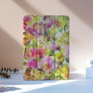May include: A tablet case with a floral watercolour design in pink, yellow, and green. The case is rectangular and stands upright on a white surface. The background is a soft peach colour. The design features flowers and leaves.
