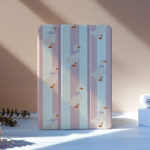 May include: A rectangular tablet case with a vertical striped pattern in pink and light blue. The case is decorated with illustrations of white geese wearing purple bows. The case is standing upright on a white surface.
