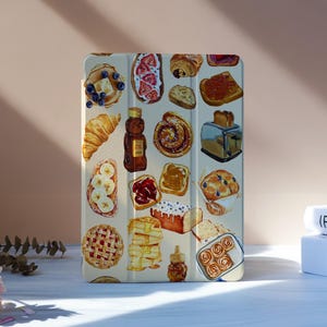 Può includere: Una custodia per tablet color crema con un'illustrazione ad acquerello di vari alimenti per la colazione. Il design include pancake, toast, pasticcini e una bottiglia di miele. Sfondo neutro con fiori.