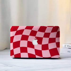 Puede incluir: Una funda para portátil a cuadros rojos y rosas con el logotipo de Apple en el centro. La funda tiene un patrón ondulado y está diseñada para un portátil. El fondo es de color claro.