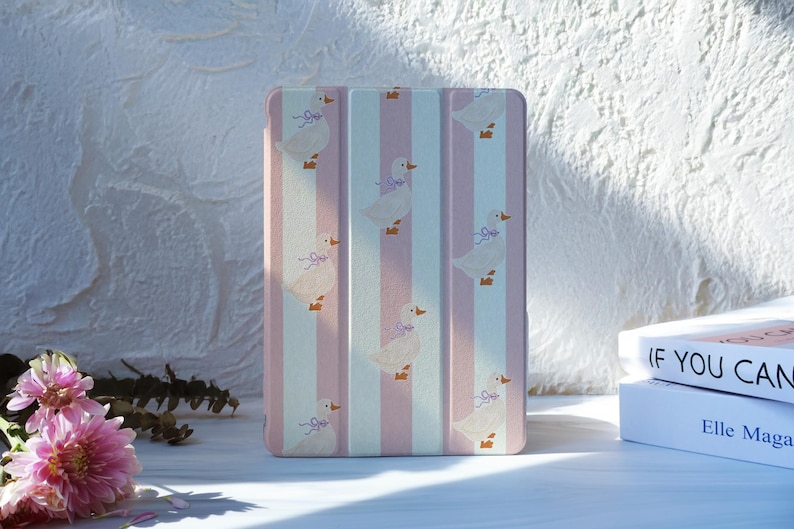 May include: A rectangular tablet case with a vertical striped pattern in pink and white. The case features a repeating pattern of white ducks with purple bows. The case is on a white surface.