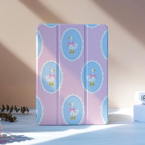 Può includere: Una custodia per tablet a righe rosa e bianche con un motivo di oche bianche con fiocchi rosa all'interno di cornici ovali blu. La custodia è in piedi su una superficie bianca, con fiori e libri sullo sfondo.