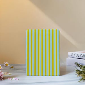 May include: A rectangular case featuring a vertical striped design in light blue and yellow. The case stands upright on a white surface, set against a soft yellow backdrop.