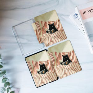 May include: Two rectangular e-reader cases and one clear plastic case cover featuring an illustration of a black cat reading a book while tucked into bed with red and white striped pillows and blankets. The cases have a smooth, flat surface.